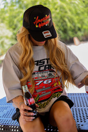 Model wearing a black trucker hat with American Woman text in red and t-shirt with racing design, holding a bottle outdoors, frontal view