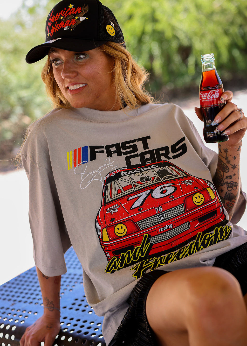 Model wearing a black trucker hat with a grey short sleeve tee with FAST CARS text and a red racing truck, sitting on a bench
