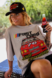 Model wearing a black trucker hat with a grey short sleeve tee with FAST CARS text and a red racing truck, sitting on a bench