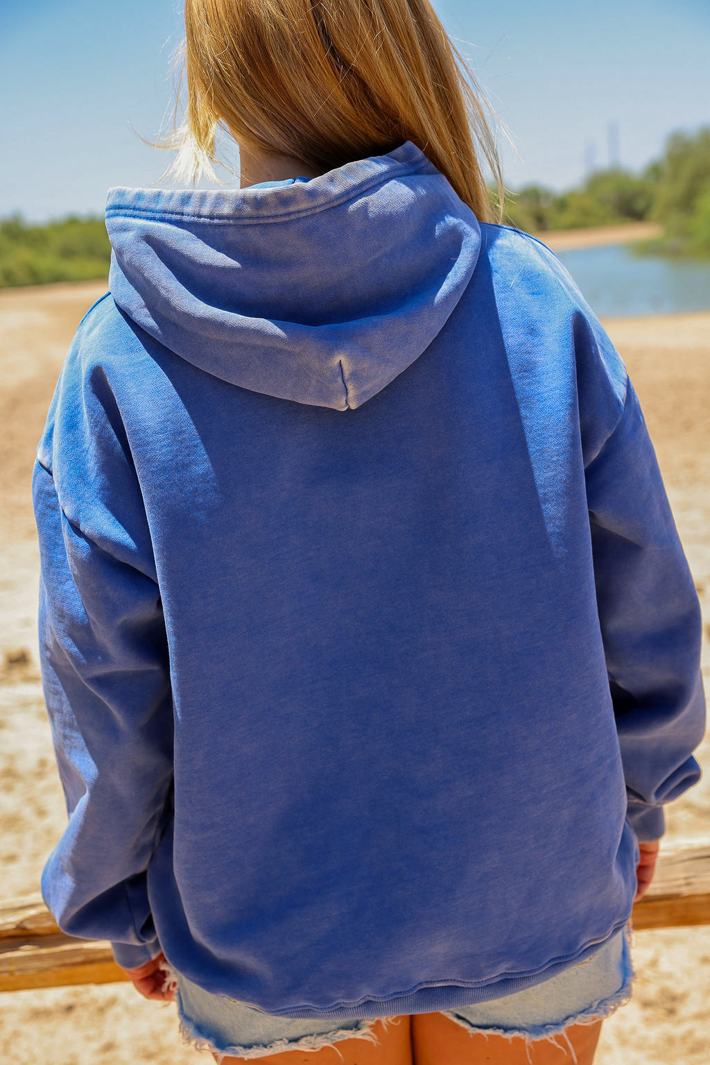 Person wearing a blue hoodie standing on a beach with water and sky in the background, close up back view