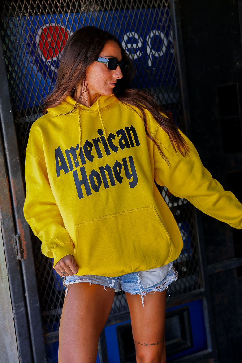 Person wearing a yellow hoodie with 'American Honey' text, standing in front of a Pepsi sign, frontal view