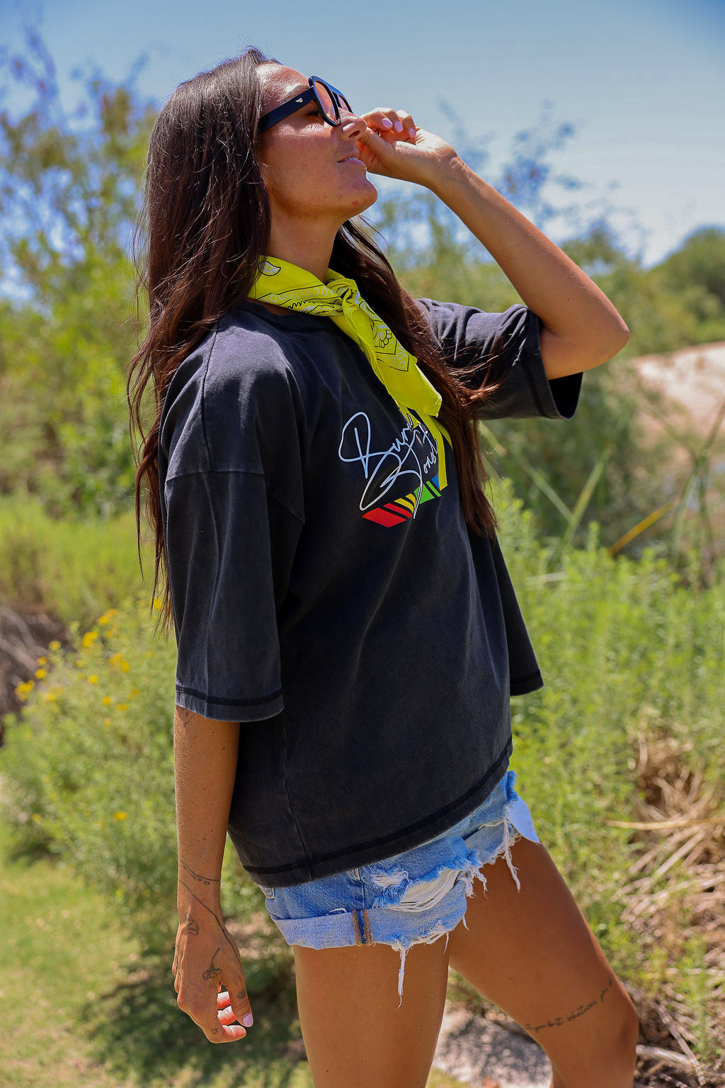 Woman wearing a black t-shirt with a colorful logo, sunglasses, and denim shorts in a natural setting, side view