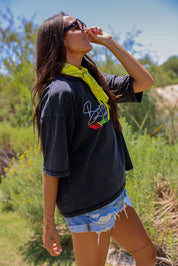 Woman wearing a black t-shirt with a colorful logo, sunglasses, and denim shorts in a natural setting, side view