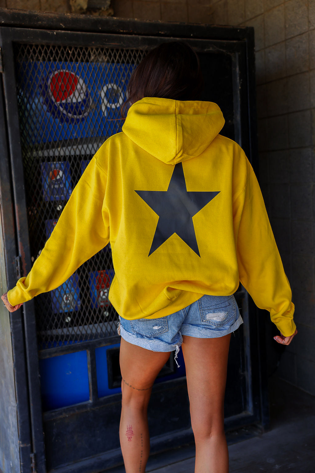 Person wearing a yellow hoodie with a black star on the back, standing in front of a Pepsi machine, black view