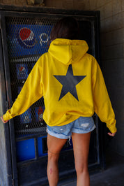 Person wearing a yellow hoodie with a black star on the back, standing in front of a Pepsi machine, black view