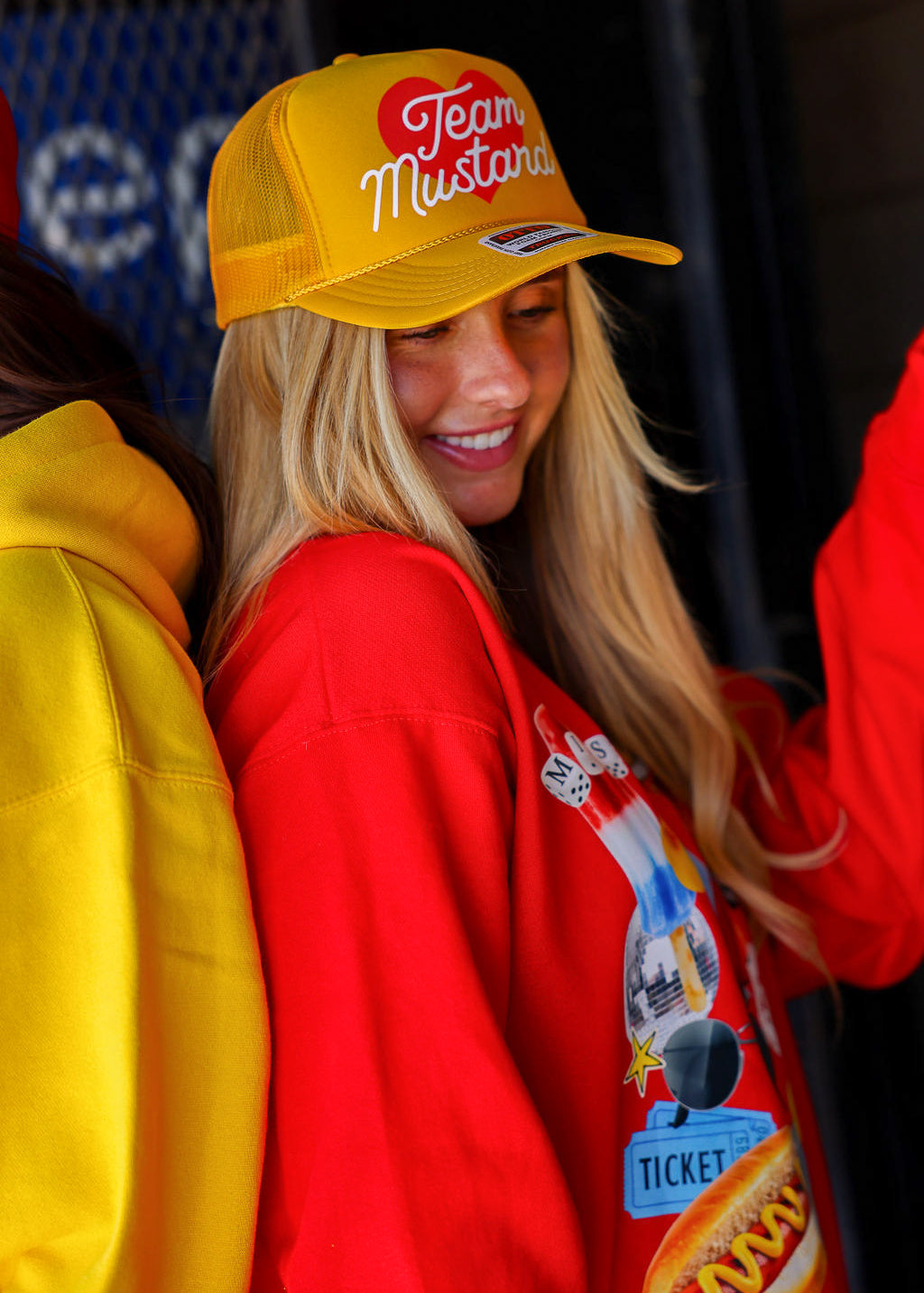 Person wearing a yellow trucker hat with 'Team Mustard' text and red crewneck with graphic design.