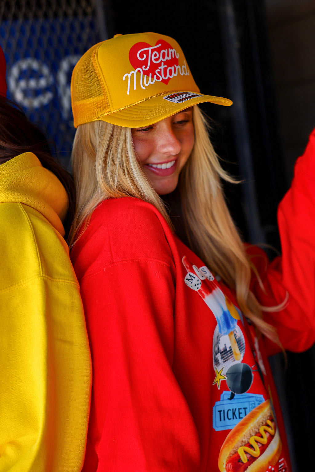 Person wearing a yellow trucker hat with 'Team Mustard' text and red crewneck with graphic design.