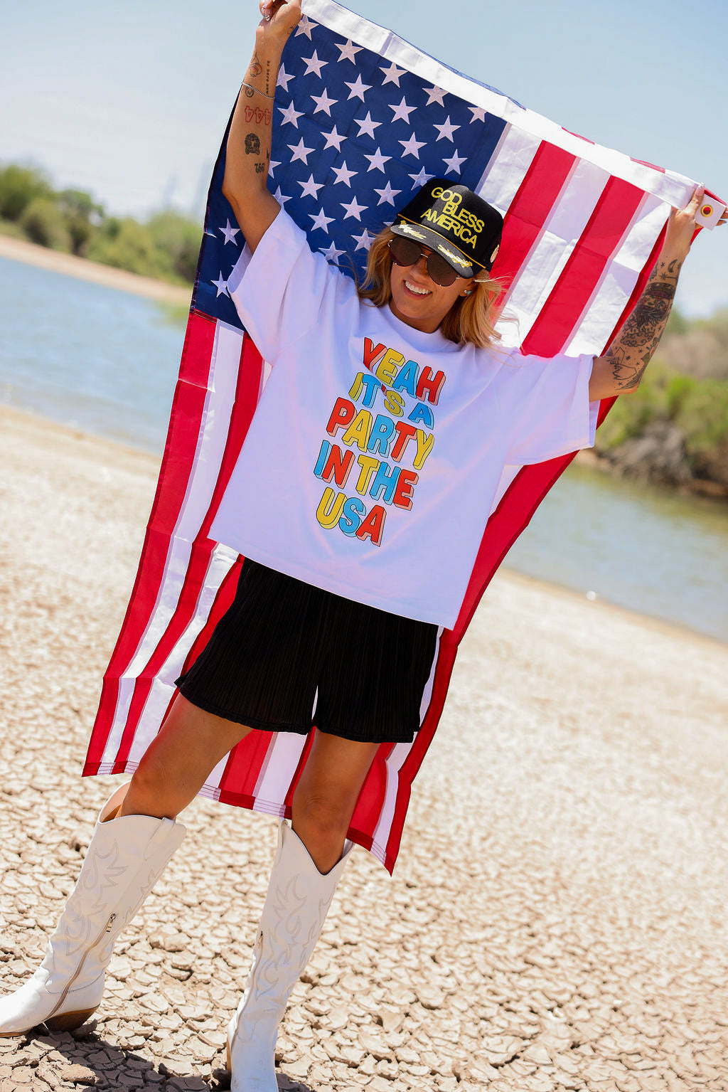 Person holding an American flag with a colorful text shirt by a body of water, frontal view