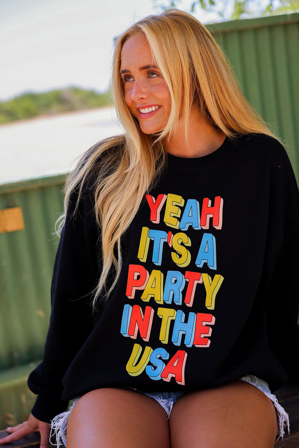 Woman wearing a black crewneck with colorful text 'Yeah It's a Party in the USA' outdoors, close up frontal view