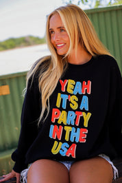 Woman wearing a black crewneck with colorful text 'Yeah It's a Party in the USA' outdoors, close up frontal view