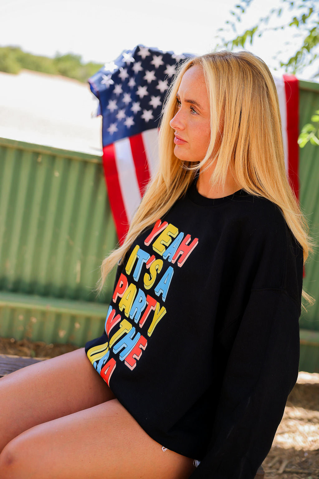 Woman wearing a black crewneck with colorful text, sitting outdoors with an American flag in the background, side view