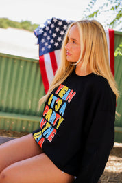 Woman wearing a black crewneck with colorful text, sitting outdoors with an American flag in the background, side view