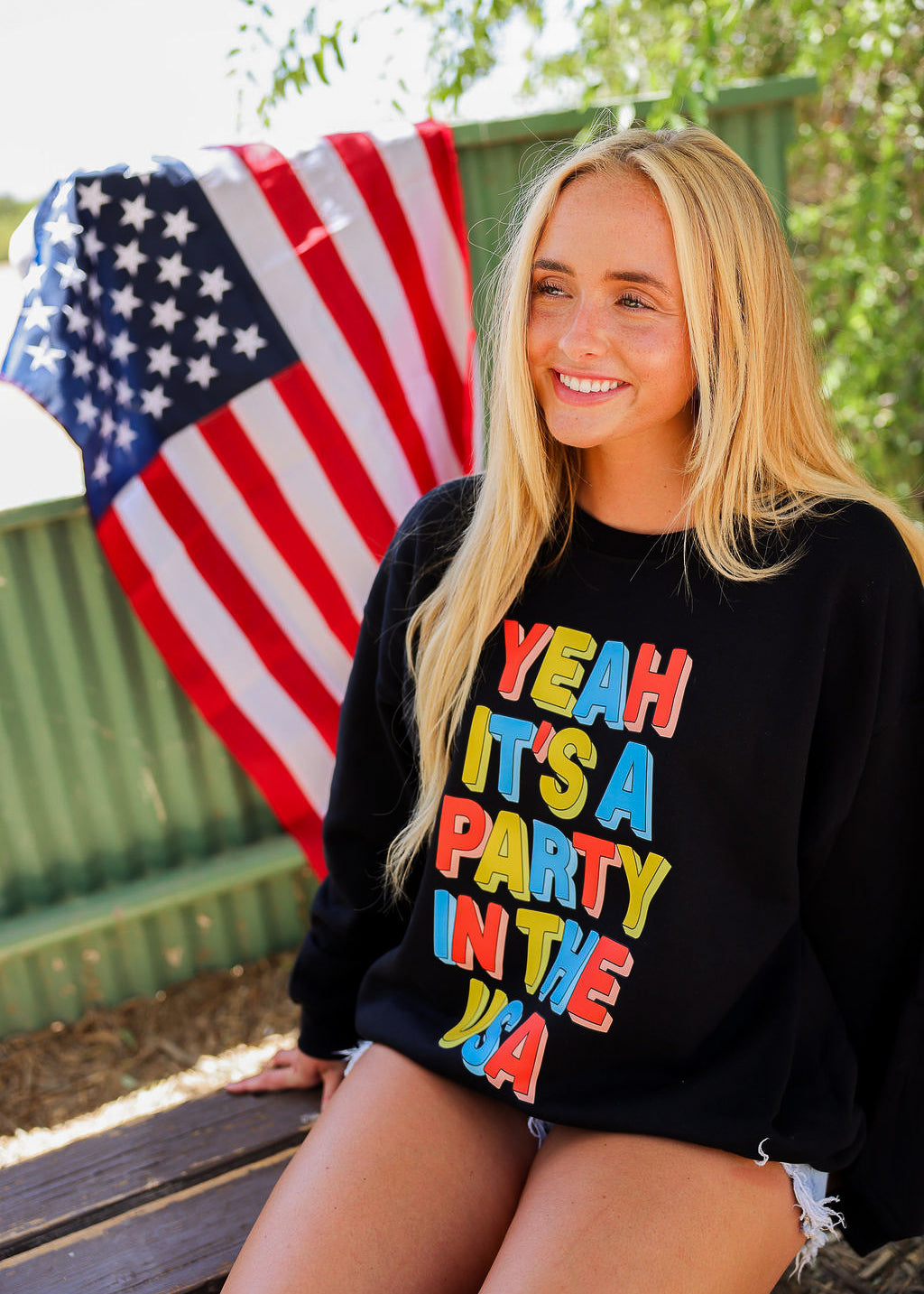 Woman wearing a black crewneck with colorful text, sitting outdoors with an American flag in the background, frontal view