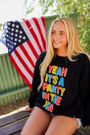 Woman wearing a black crewneck with colorful text, sitting outdoors with an American flag in the background, frontal view