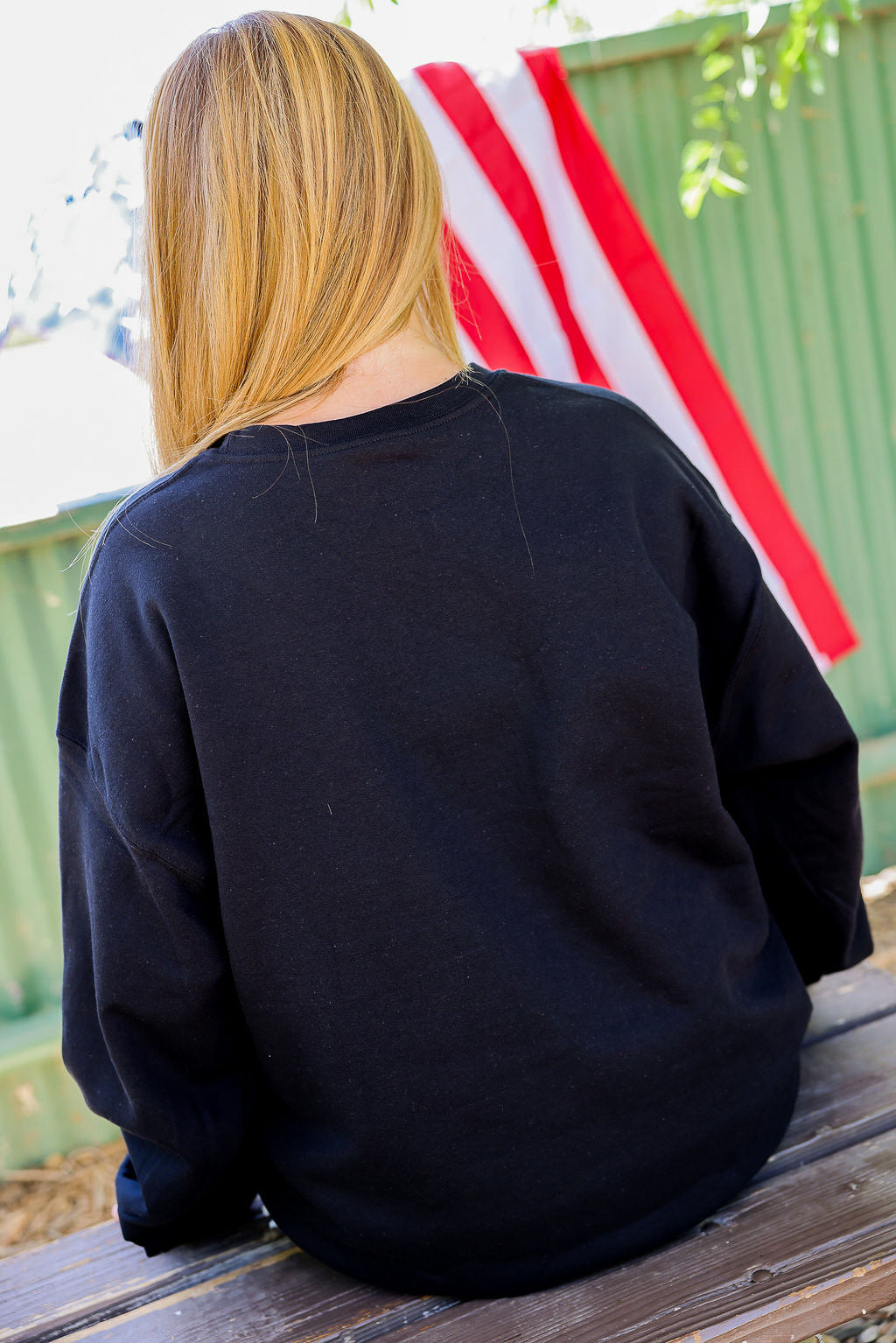 Person wearing a black crewneck sitting on a wooden bench with an American flag in the background, back view