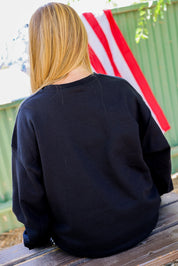 Person wearing a black crewneck sitting on a wooden bench with an American flag in the background, back view