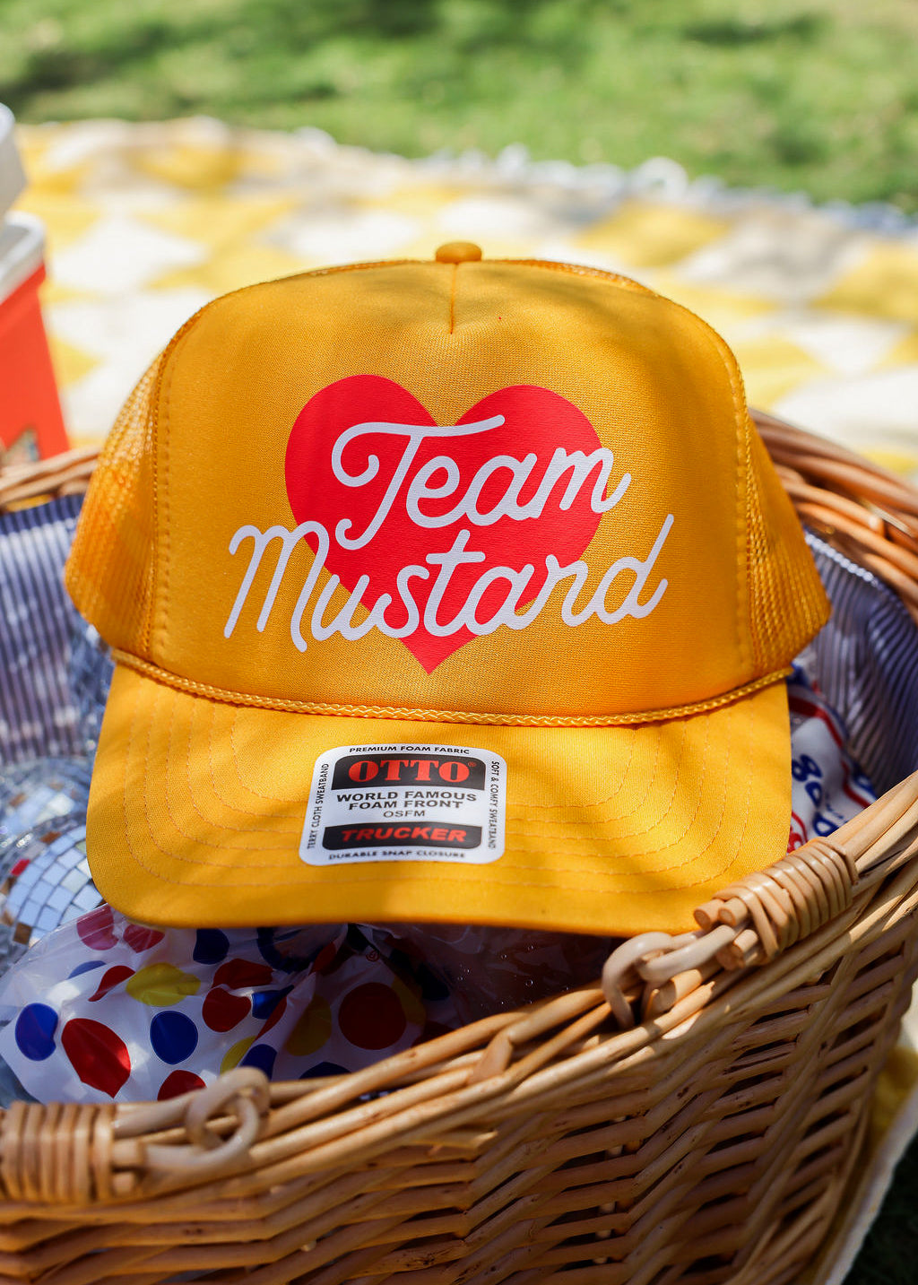 Yellow trucker at with 'Team Mustard' text and red heart in a wicker basket outdoors, frontal view