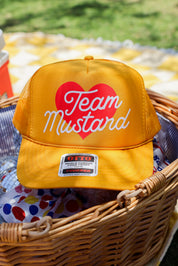 Yellow trucker at with 'Team Mustard' text and red heart in a wicker basket outdoors, frontal view
