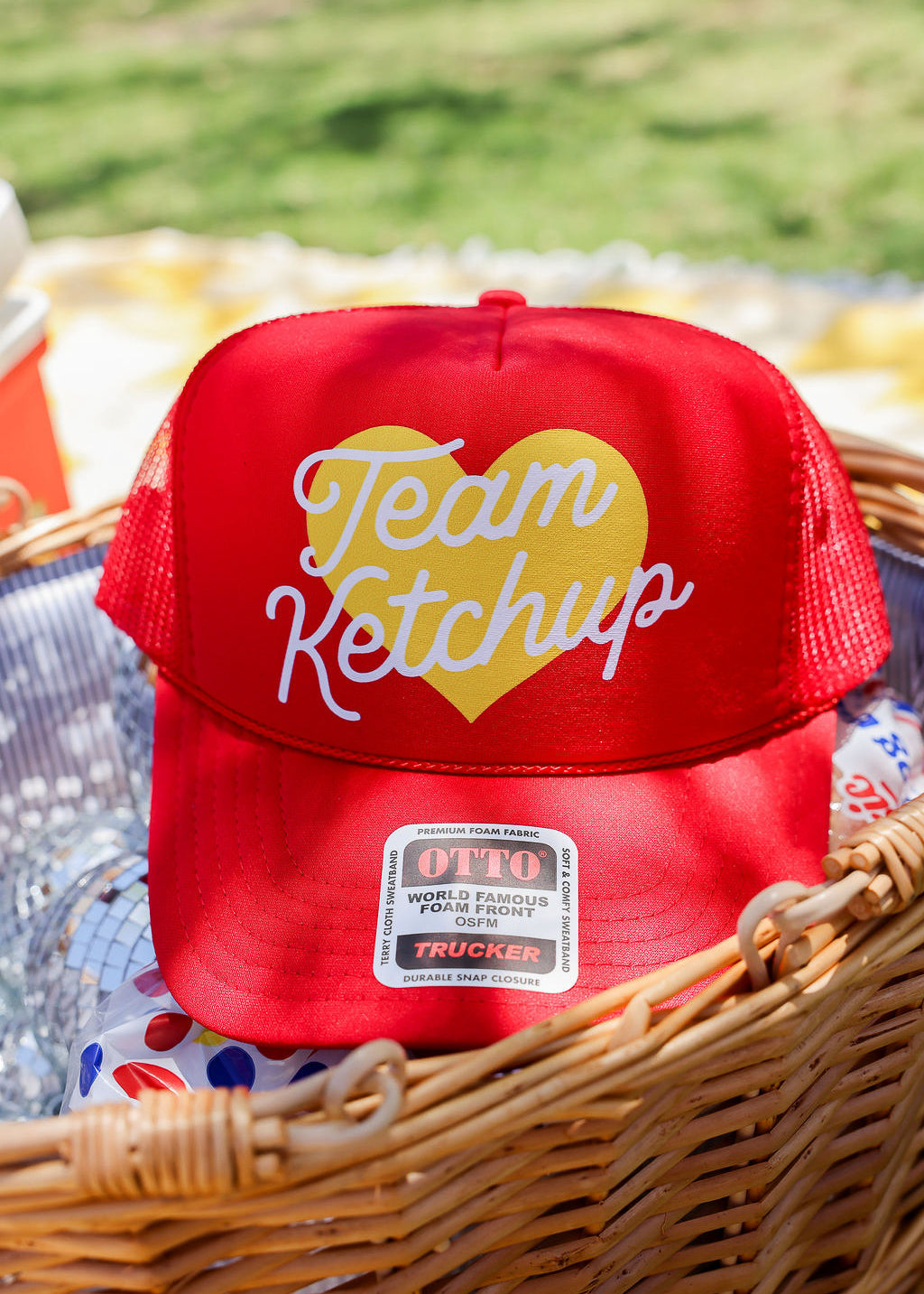 Red truker hat with 'Team Ketchup' text in a wicker basket outdoors, frontal view