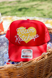 Red truker hat with 'Team Ketchup' text in a wicker basket outdoors, frontal view