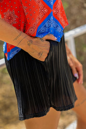 Model wearing blue and red bandana top, with hand in pocket of metallic black pleated shorts blurred natural background, close up side view
