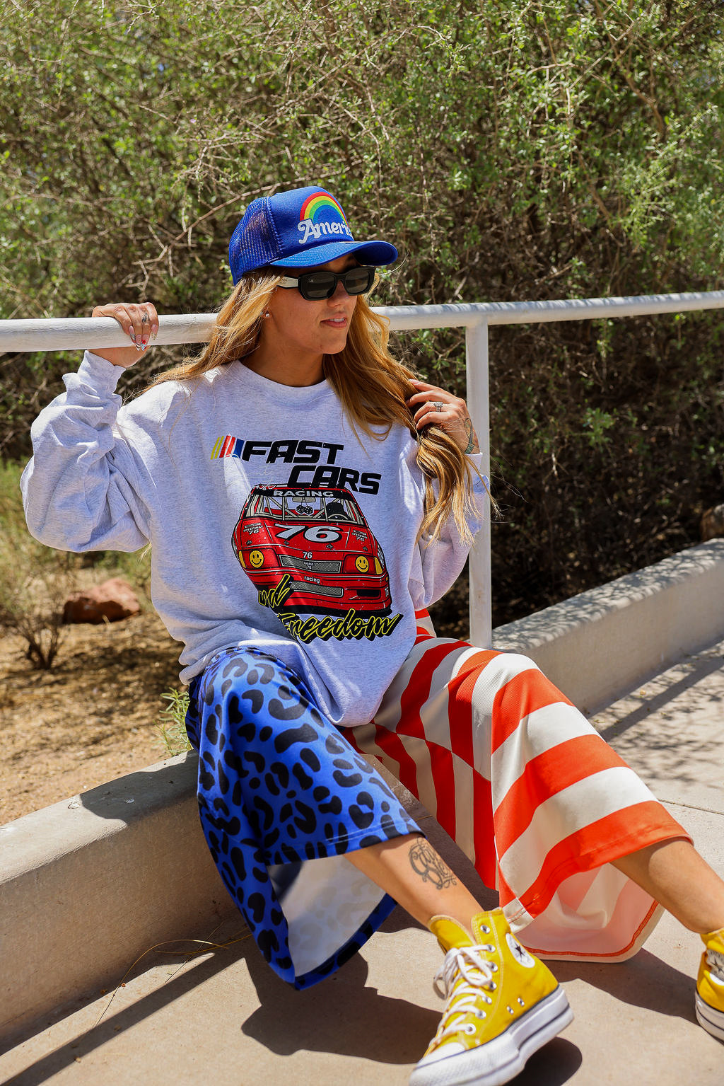 Model wearing a grey sweatshirt with a car design, blue trucker hat, and colorful pants sitting outdoors, frontal view