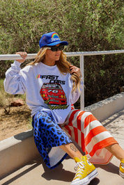 Model wearing a grey sweatshirt with a car design, blue trucker hat, and colorful pants sitting outdoors, frontal view