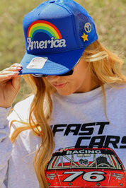 Model wearing a blue trucker hat with 'America' and rainbow design, and a grey crewneck with racing car graphic, close up frontal view