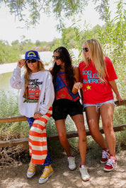 Three women posing outdoors by a lake, wearing colorful clothing and accessories, frontal view