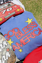 Blue folded t-shirt with 'GOD BLESS AMERICA' text and stars, placed on grass.
