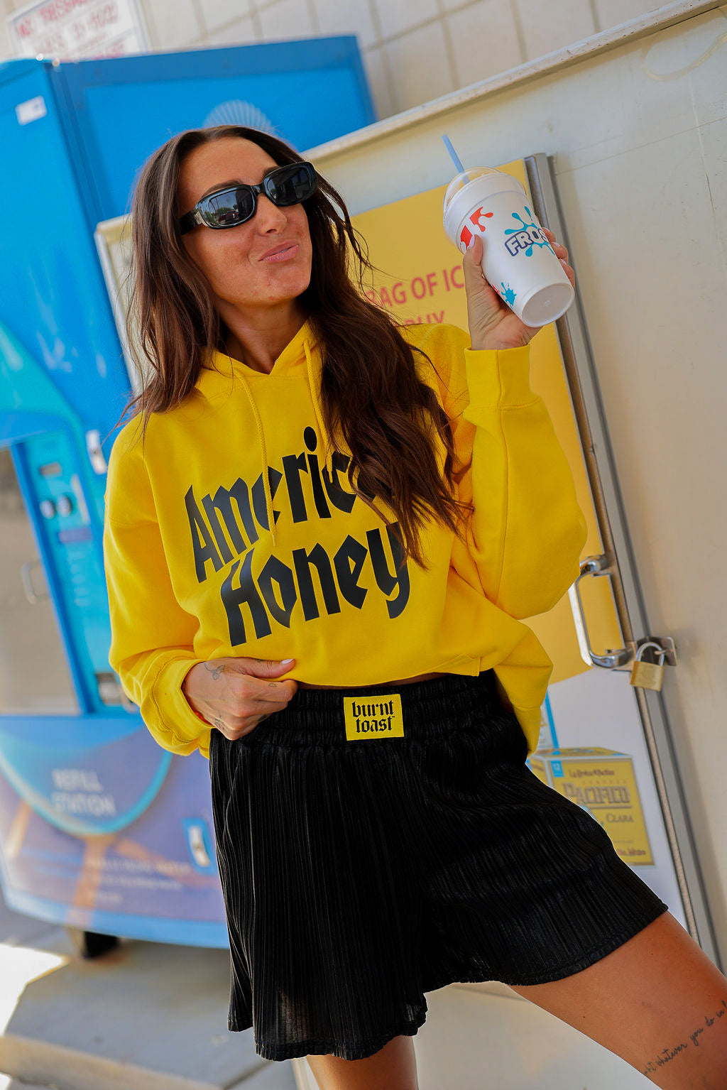 Person wearing a yellow 'America Honey' hoodie and black plisse shorts with yellow burnt toast label on waistband, sunglasses, holding a cup.