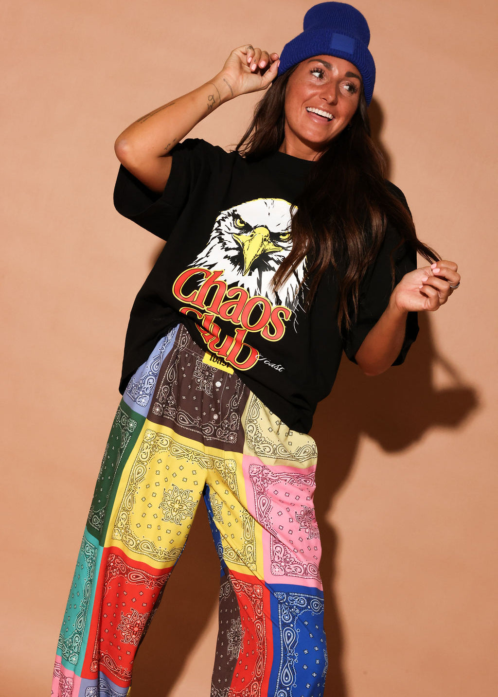 Model wearing a black t-shirt with an eagle graphic and colorful bandana-patterned pants on a beige background, frontal view