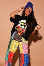 Model wearing a black t-shirt with an eagle graphic and colorful bandana-patterned pants on a beige background, frontal view