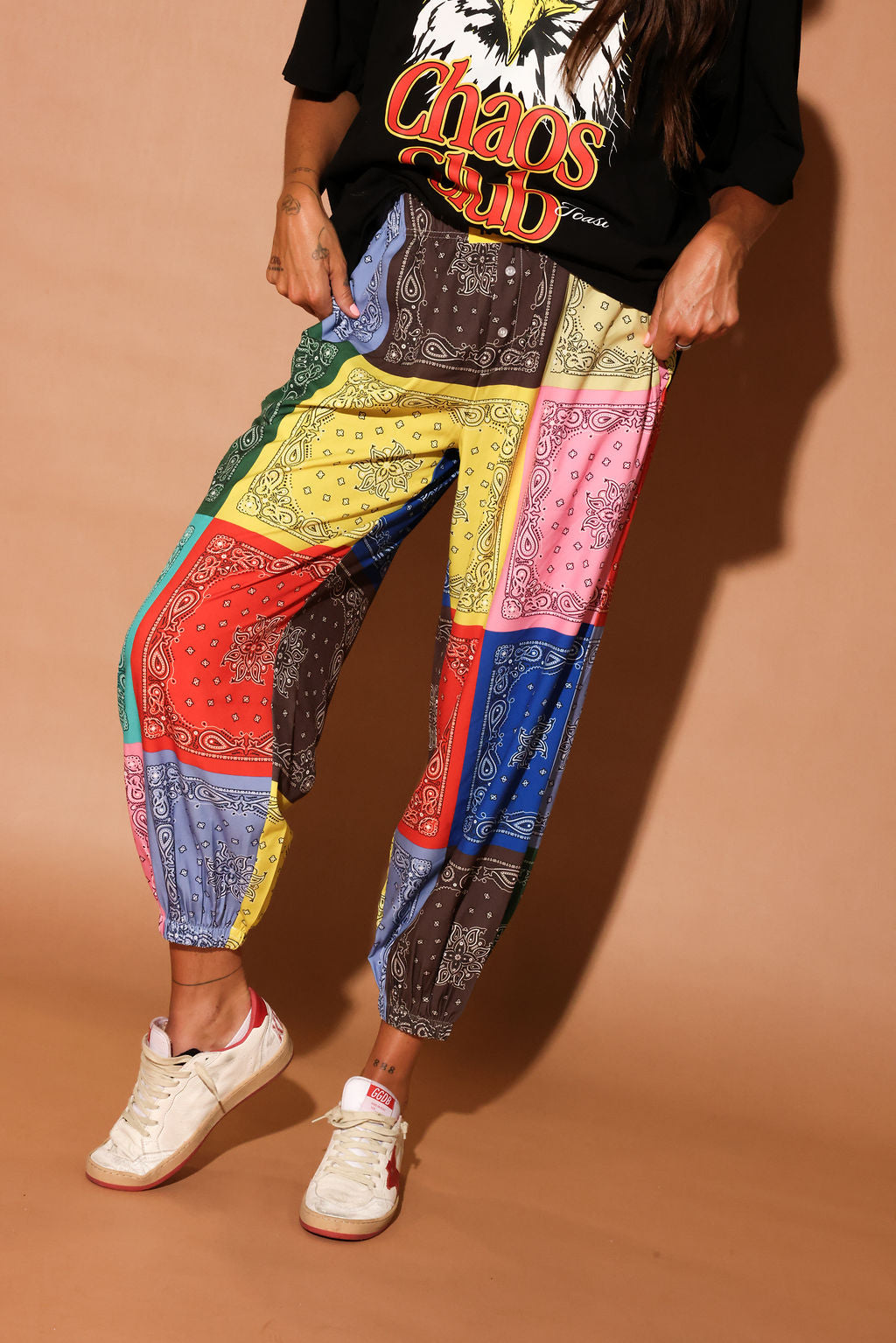 Person wearing colorful bandana patterned pants and a black t-shirt with 'Chaos' text on a brown background, bottom half of body view