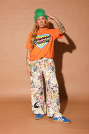 Person wearing an orange t-shirt with text, colorful paint splatter pants, and a green beanie on a brown background, frontal view