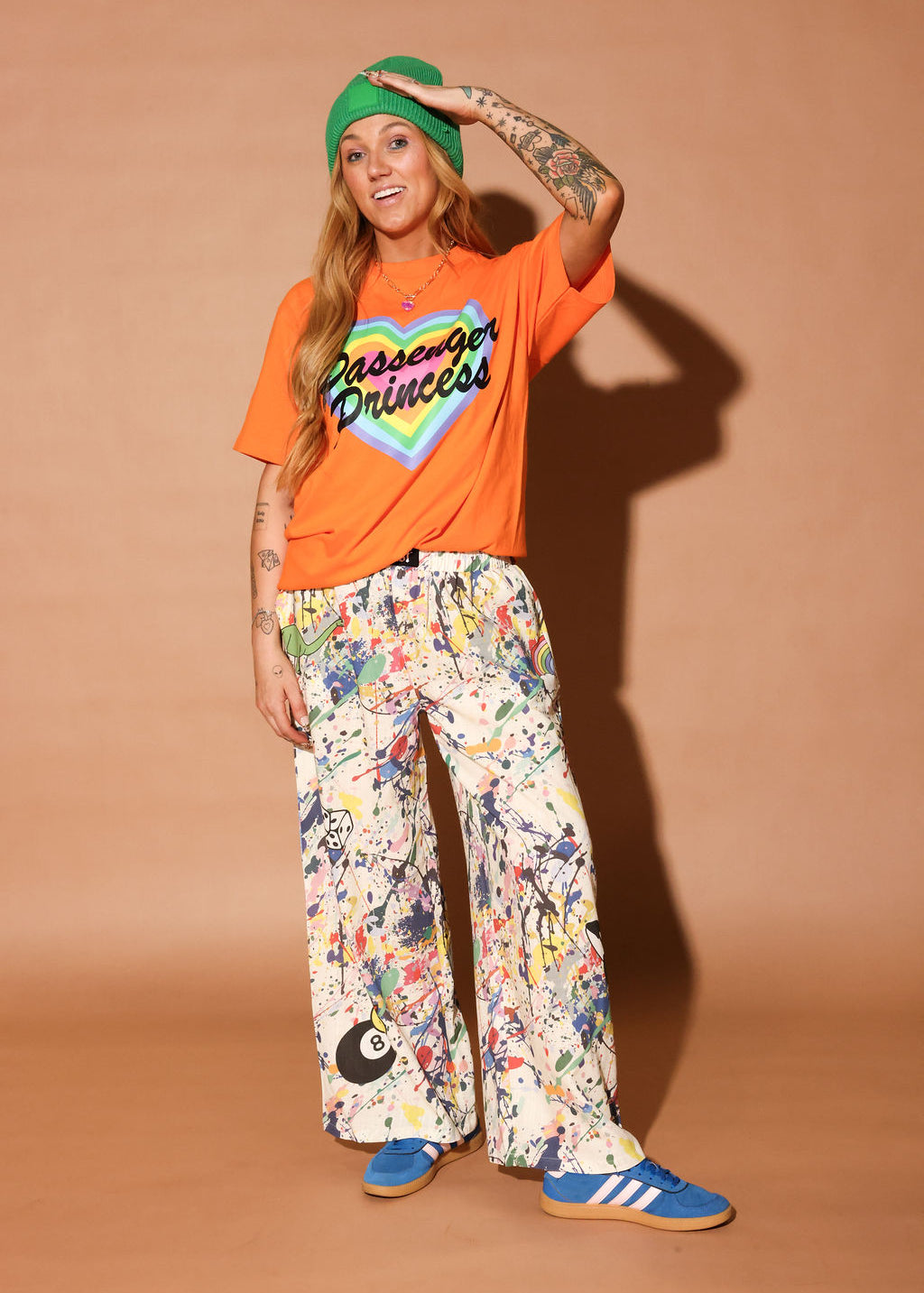 Person wearing an orange 'Passenger Princess' t-shirt, colorful pants, and a green beanie on a brown background, frontal view