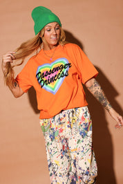 Person wearing an orange 'Passenger Princess' shirt with colorful pants and a greenie beanie against a beige background, frontal view