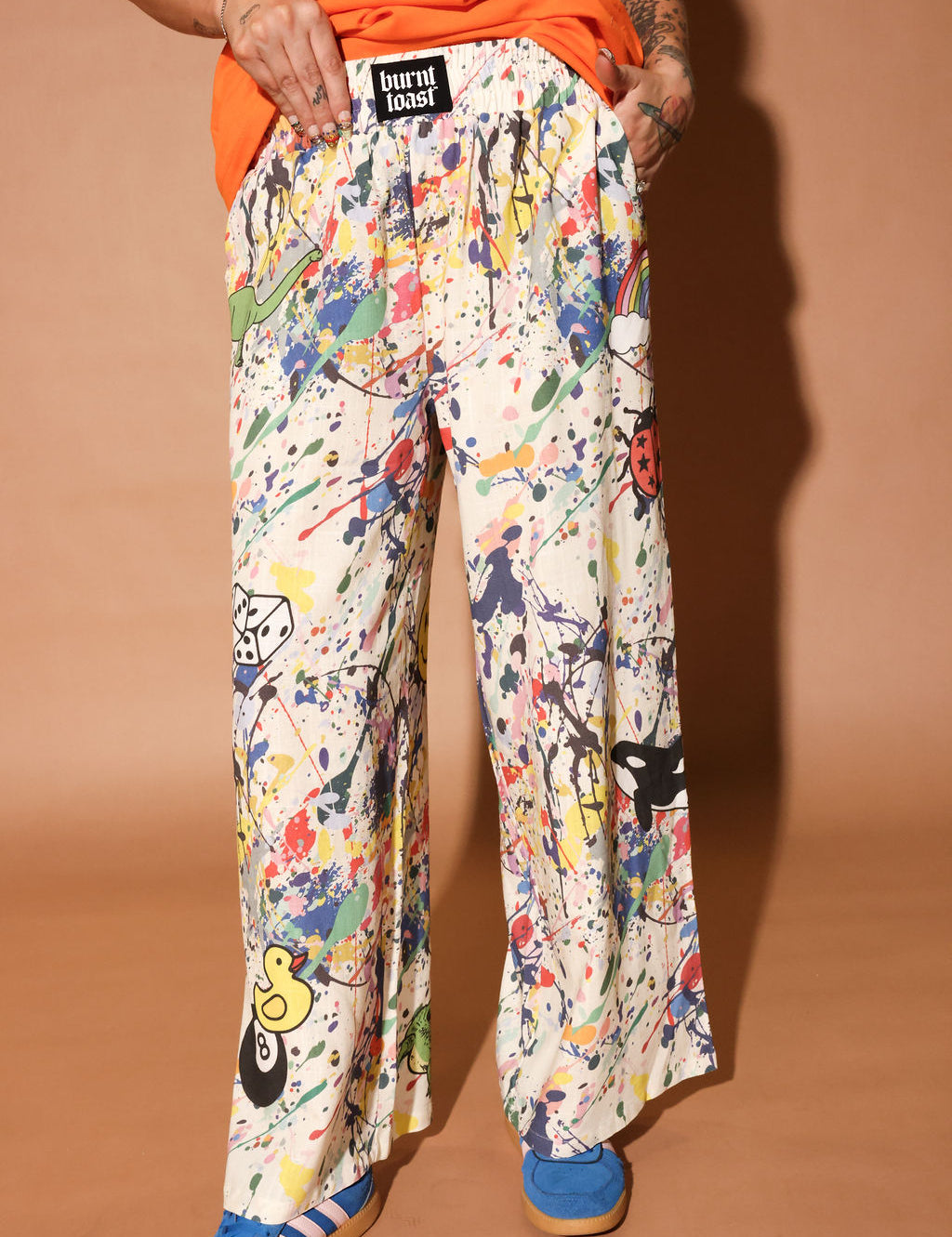 Person wearing colorful patterned pants with paint splatters and a brand label on a brown background, close up view