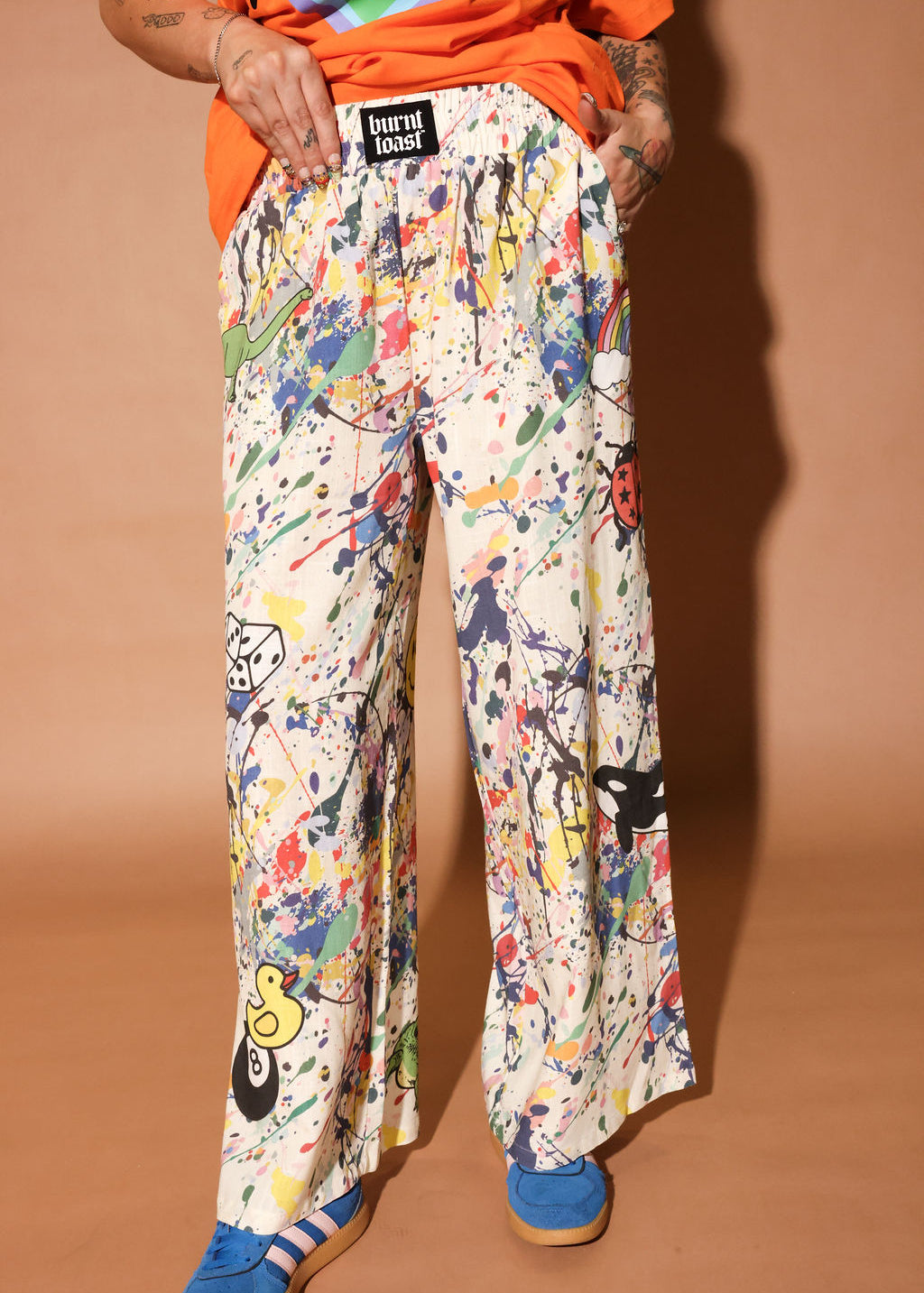 Person wearing colorful patterned pants with paint splatters and a brand label on a brown background, close up view
