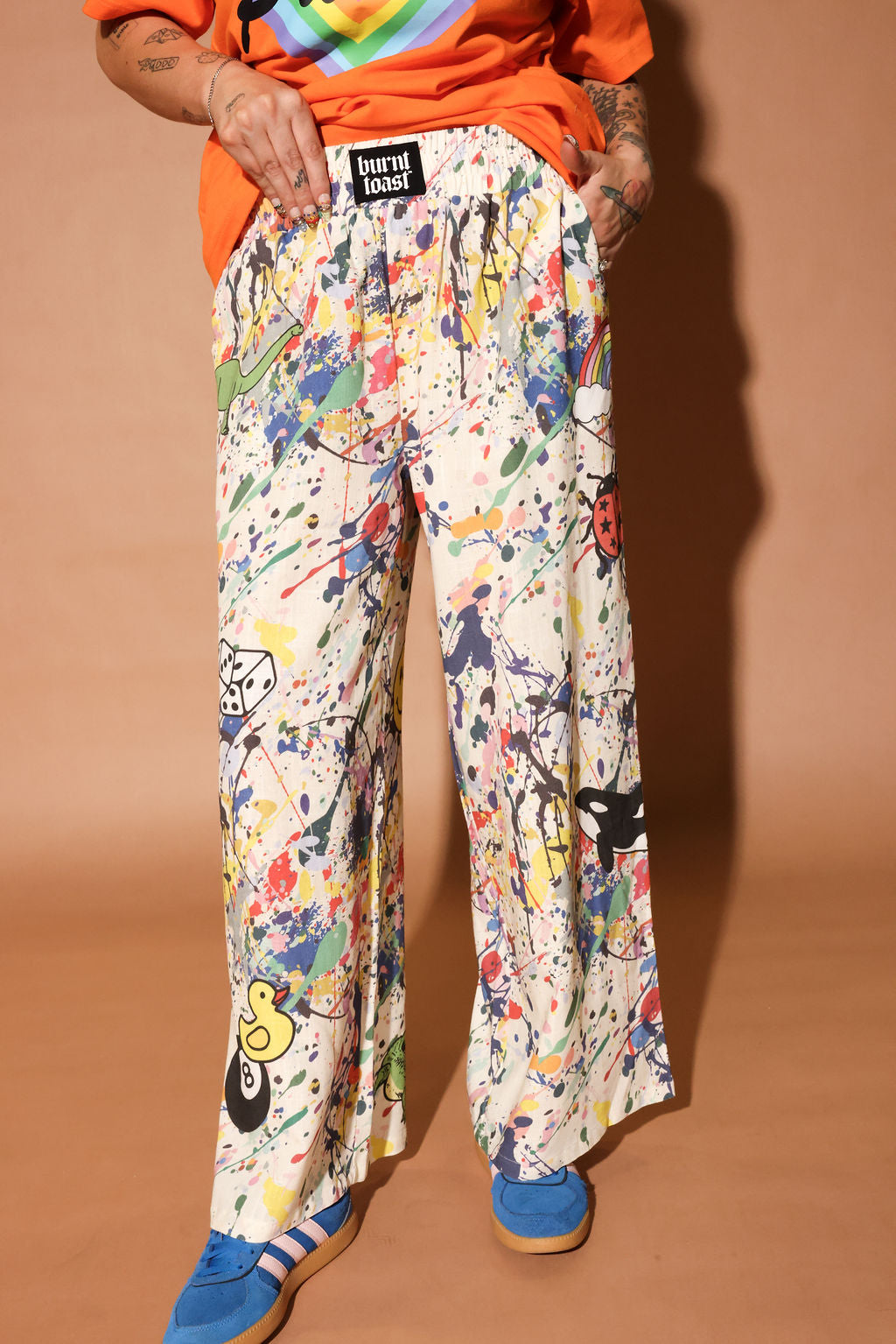 Person wearing colorful patterned pants with paint splatters and a brand label on a brown background, close up view