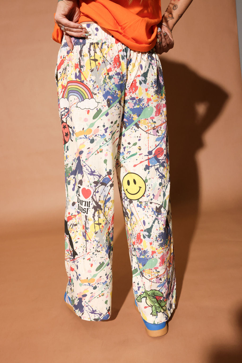 Person wearing colorful patterned pants with paint splatters and a smiley face on a brown background, back view