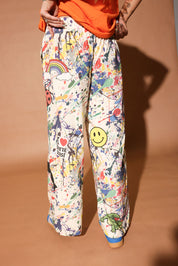 Person wearing colorful patterned pants with paint splatters and a smiley face on a brown background, back view