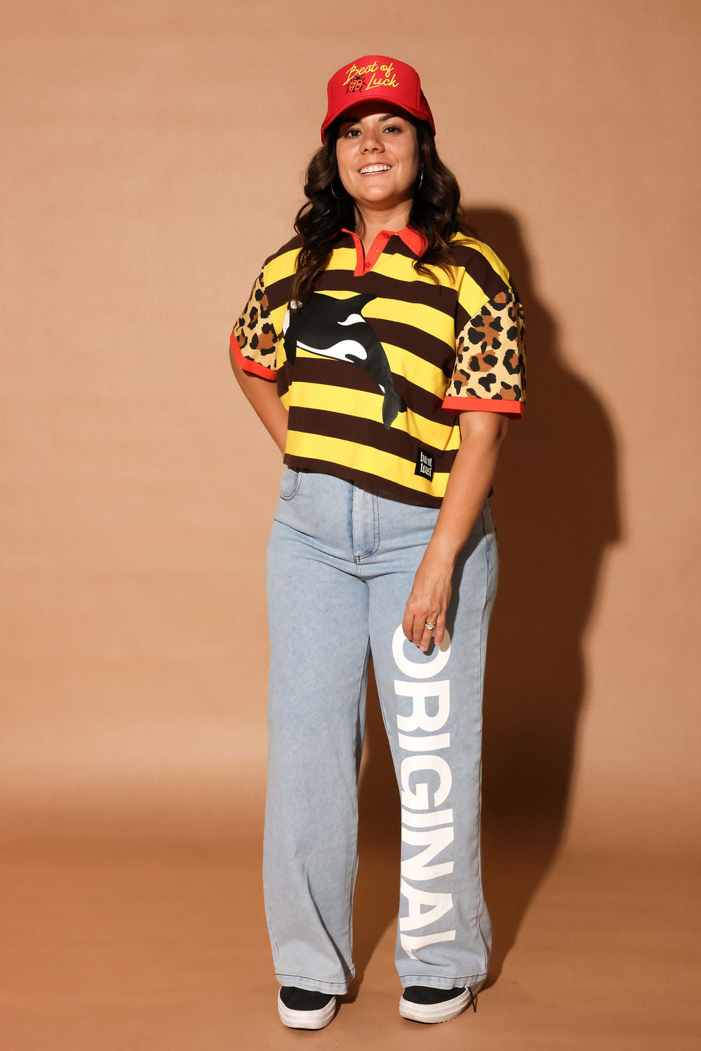 Model wearing a yellow and brown striped polo with an orca whale design and 'burnt toast' logo, denim jeans and a red trucker hat, on a brown background, frontal view
