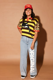 Model wearing a yellow and brown striped polo with an orca whale design and 'burnt toast' logo, denim jeans and a red trucker hat, on a brown background, frontal view