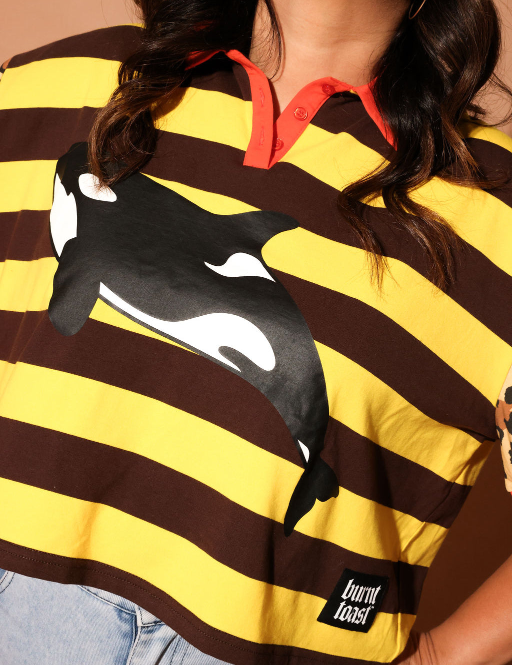 Model wearing a yellow and brown striped polo with an orca whale design and 'burnt toast' logo, close up frontal view