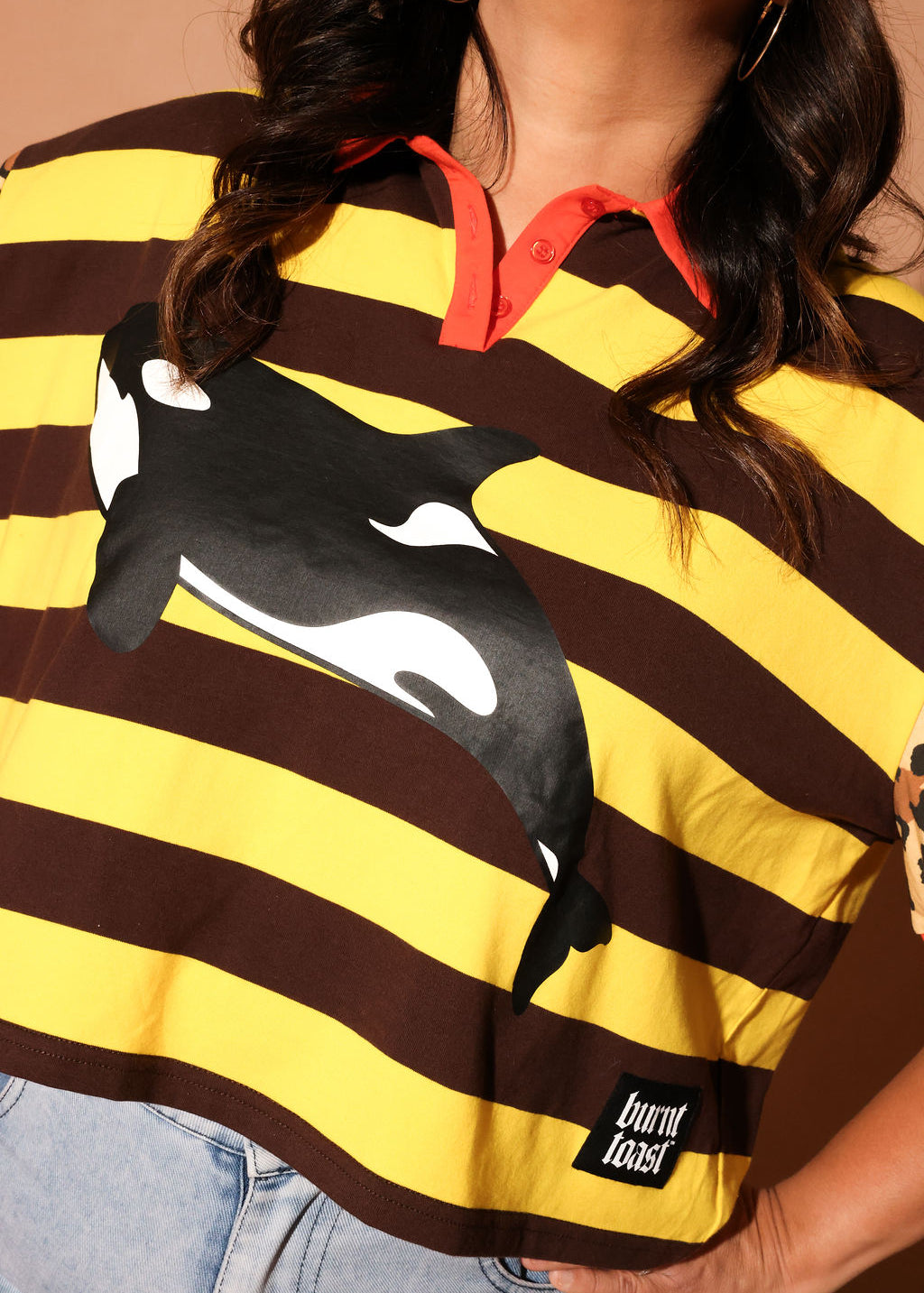Model wearing a yellow and brown striped polo with an orca whale design and 'burnt toast' logo, close up frontal view