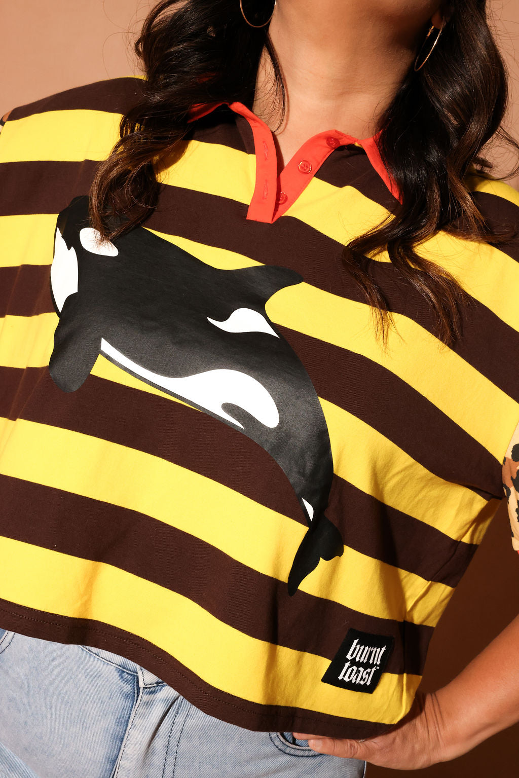 Model wearing a yellow and brown striped polo with an orca whale design and 'burnt toast' logo, close up frontal view