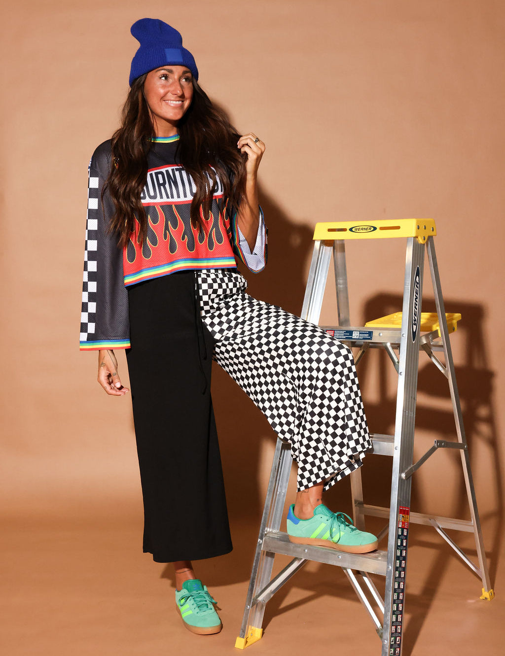 Model wearing a color jersey with flames, and black and white checkered cropped pants, black beanie, standing in front of a building, frontal view
