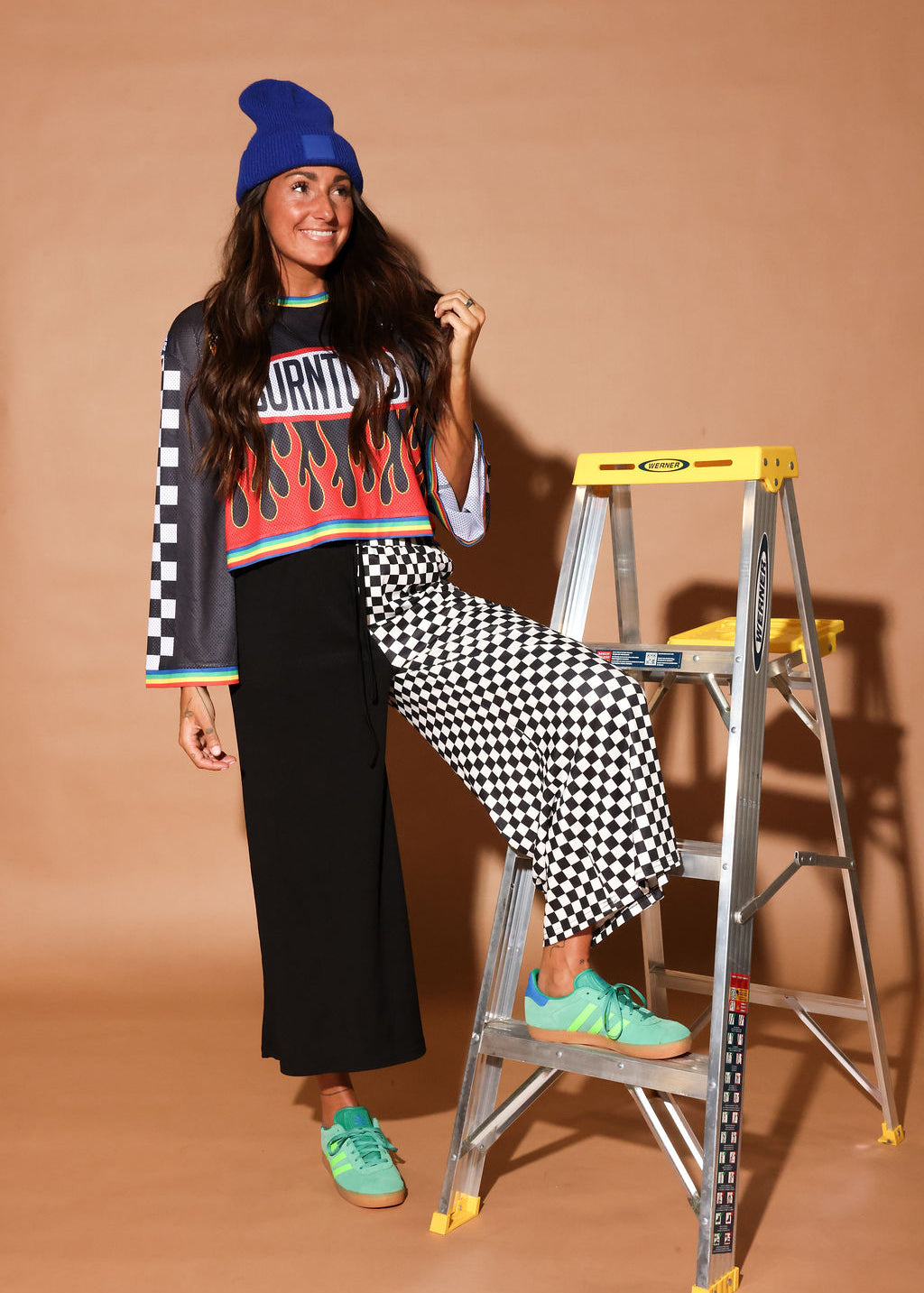 Model wearing a color jersey with flames, and black and white checkered cropped pants, black beanie, standing in front of a building, frontal view
