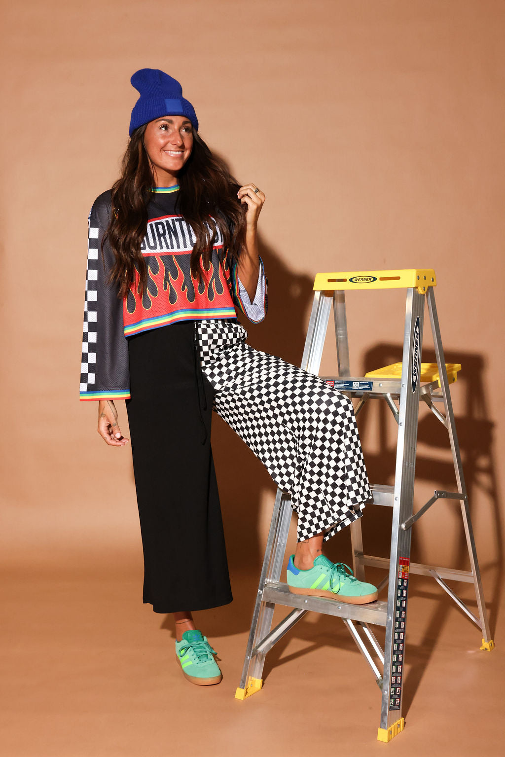 Model wearing a color jersey with flames, and black and white checkered cropped pants, black beanie, standing in front of a building, frontal view